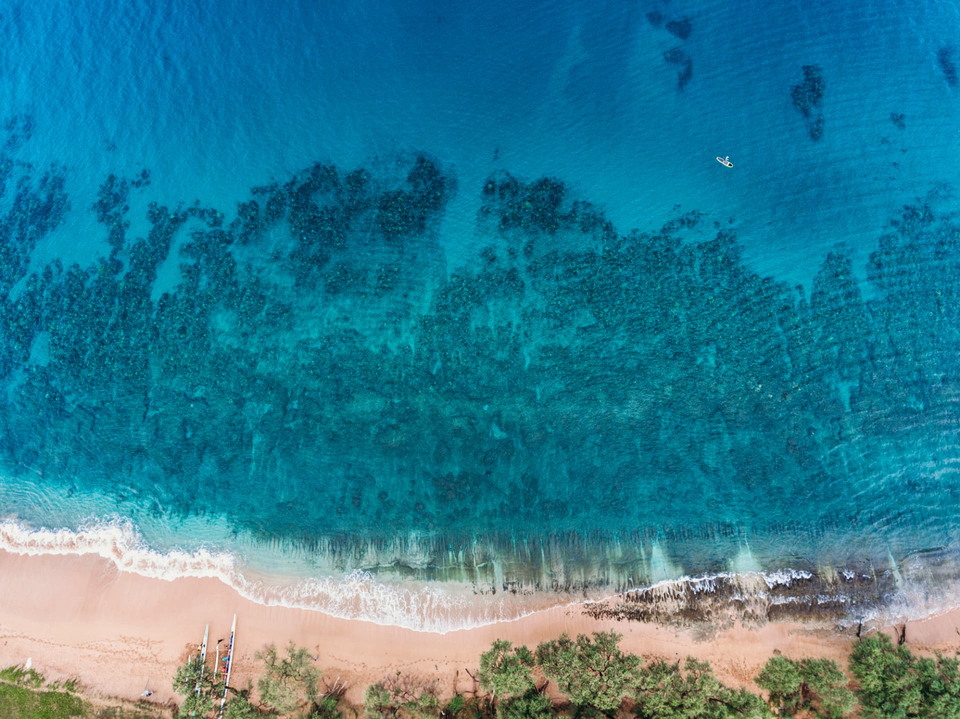 Aerial view of deep blue water over a wide reef with a single paddler