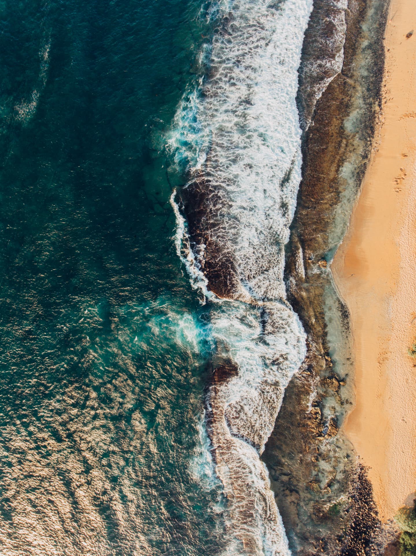 Aerial view of surf meeting a rocky shoreline and pale sand