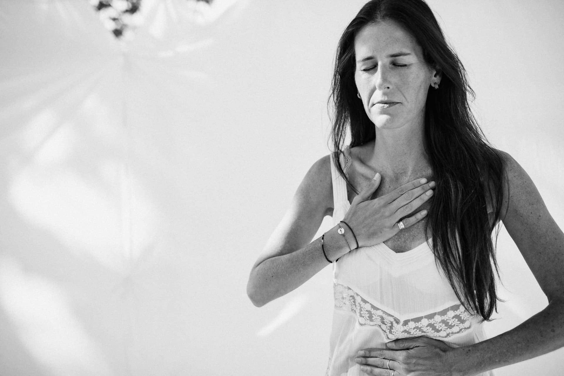 A woman standing with eyes closed, one hand resting on her heart and the other on her belly